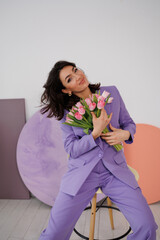 Beautiful brunette woman in violet costume with bucket of tulips, flowers, sitting on high chair. Girl smile, happy. Portrait of young woman. Long legs, barefoot