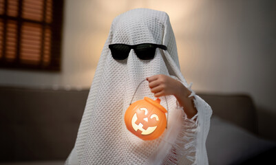Little children wearing a white scary ghost costume holds a bucket of pumpkins for Halloween.