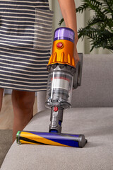 Beautiful slender caucasian girl in a striped dress is vacuuming the sofa upholstery with a modern wireless vertical vacuum cleaner with a special nozzle for fabric. - 651247898
