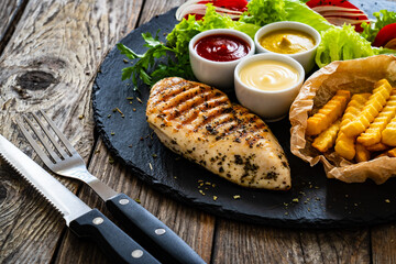 Grilled chicken breast, French fries and fresh vegetables on wooden table
