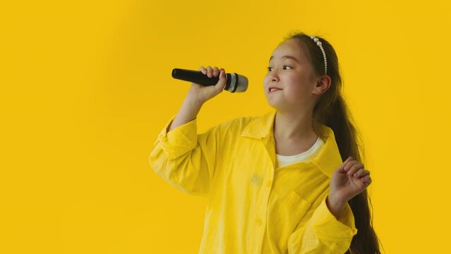 Young asian little girl passionately singing a song to the microphone and smiling on a yellow background. Talented child showing her creative skills