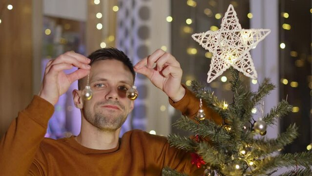 Middle aged man holds two glass Christmas balls in hands and looks at them indifferently. Lonely hispanic sad guy not happy about holidays, puts decorative toy in mouth as sign of joyful protest.