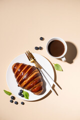 Freshly baked croissant with chocolate on a white plate and blueberries on a yellow background with cup of black coffee. The concept of a delicious homemade breakfast.
