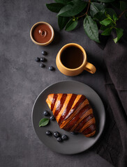 Freshly baked chocolate croissant with blueberries on a gray plate on a dark background with a cup of black coffee. The concept of a delicious homemade breakfast in rustic style.
