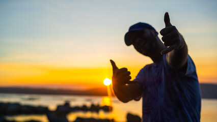 young man wearing a cap doing a surfer greeting to the camera at the sunset