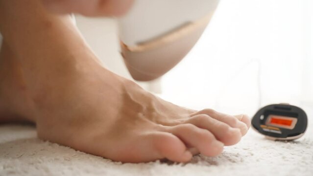 A home photo epilator with various attachments, a woman removing hair from her foot, close-up shot, on a white soft carpet in a bright room.