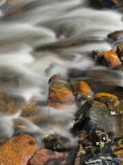 Long exposure river bed. Smooth water as flows over pebbles in a rivers bed, browns and green pebbles with smooth water around. On a summers sunny day in the forest.
