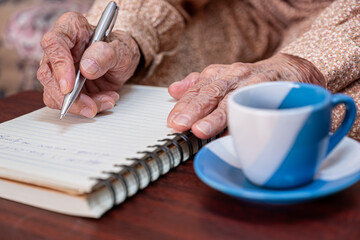 wrinkled hands for elderly person writing notes on his note book while drinking coffee