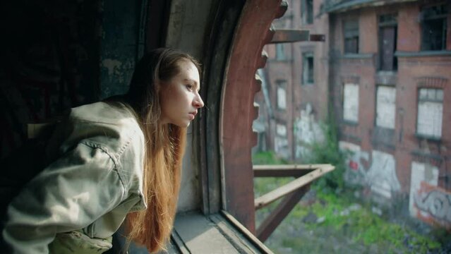 A young caucasian beautiful woman with long hair in green jacket looks thoughtfully through an antique window frame without glass at an old red abondoned destroyed building at sunny day