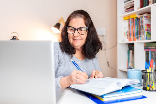 Mature Brazilian Woman Studying At A Home Office Desk, Reading With A Laptop Beside Her, Suggesting Online Learning Or Internet Research. Concept: Older Individuals Returning To Education