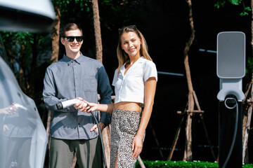 Young couple travel with EV electric car charging in green sustainable city outdoor garden in...