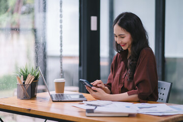 A millennial, mixed-race Asian businesswoman is using a smartphone at her desk in a modern office.