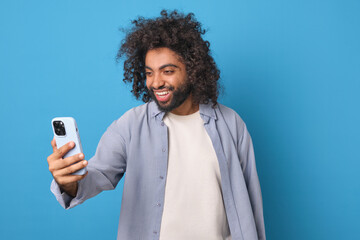 Fototapeta na wymiar Young positive attractive Arabian man in casual clothes holds phone in hand and smiles using mobile applications for video chatting and communicating with new people via internet stands in blue studio