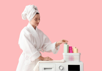 Young woman in bathrobe with laundry detergent and washing machine on pink background