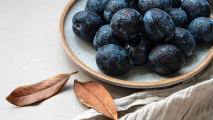 Fresh plums with water drops on plate, closeup, brown fall leaves on neutral beige table background