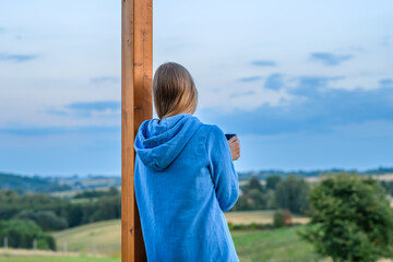 Kobieta podziwia krajobraz stojąc na ganku domku w górach z kubkiem herbaty - obrazy, fototapety, plakaty