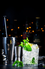 Tommy's margarita summer cocktail drink with tequila, lime juice, lemon, syrup, mint and ice in glass, dark background, bar tools and bottles