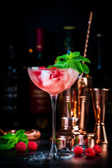 Raspberry daiquiri pink cocktail with white rum, sugar syrup, fruit juice, lime, berry and ice in glass. Black bar counter background, bar tools and bottles