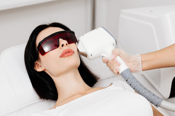 A beautiful young woman in special glasses undergoes a laser hair removal procedure on her face in...