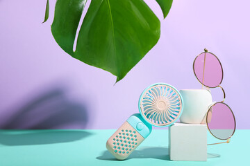 Plaster podium with portable electric fan, monstera leaf and stylish eyeglasses on color background