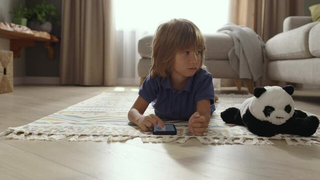 Portrait of cute smiling boy 5-7 years old lying on carpet and watching robot vacuum cleaner. Home sweet home. Smart technologies. People spending time for relaxing instead of households. 