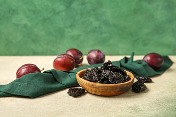 Bowl with tasty prunes and ripe plums on white grunge table against green background