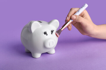 Female hand with makeup brush and piggy bank on lilac background