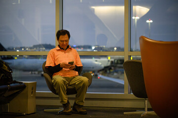 Man reading his smartphone, sitting by the window at the airport departure area. Out-of-focus airplanes parked outside the terminal window at dusk.