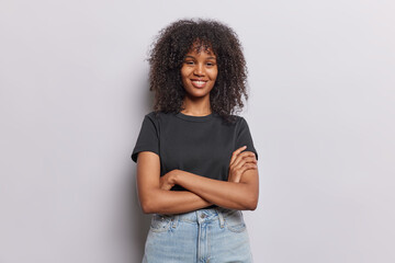 Photo of positive curly haired woman poses with arms crossed smiles gladfully dressed in casual...
