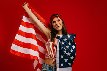 Attractive young hipster woman carrying American flag and smiling while standing on red background