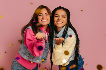 Two joyful young women blowing colorful confetti and smiling against pink background