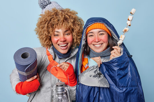Winter tourism. Studio photo of young happy smiling broadly African american and European females with red cheeks standing close to each other in warm clothes holding mat lantern and fried marshmello