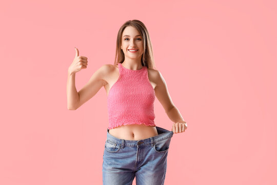 Beautiful young woman in loose jeans showing thumb-up gesture on pink background. Weight loss concept