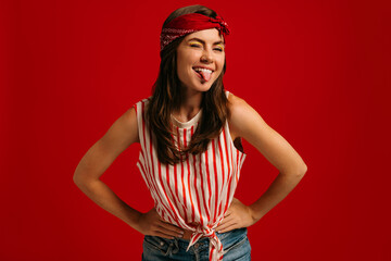 Playful young hipster woman holding hands on hip and sticking out tongue while standing on red background