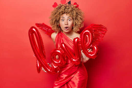 Holiday celebration. Indoor photo of young pretty surprised African american lady in red dress standing isolated in centre holding love sign seeing something unusual or unexpexted. St. Valentine's Day