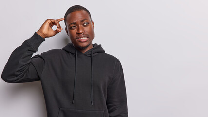 Clueless dark skinned bearded man keeps finger on head and looks away focused aside on copy space...