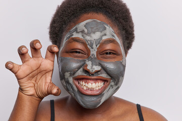 Funny chubby young African woman with curly hair makes paw while undergoes beauty procedures clenches teeth has piercing in nose isolated over white background. Wellness and skin care concept