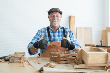 Caucasian senior old bearded man carpenter in apron, hat and gloves sitting smiling, thumbs up, working in workshop, tools machine small wooden toy model or handcrafts is on table, looking at camera