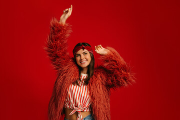 Joyful young hipster woman in fluffy coat dancing against red background
