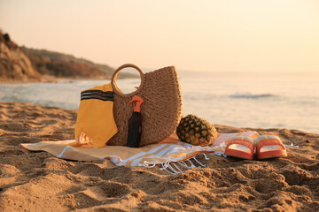 Stylish beach bag, flip-flops, pineapple, sunscreen spray, swimsuit and towel on sand near sea - obrazy, fototapety, plakaty