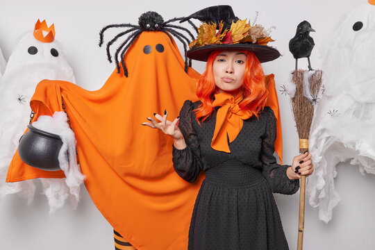 Halloween party concept. Indoor photo of young hesitant European woman wearing black dress of witch holding broom with raven on top spreading palms looking straight at camera surrounded by ghosts