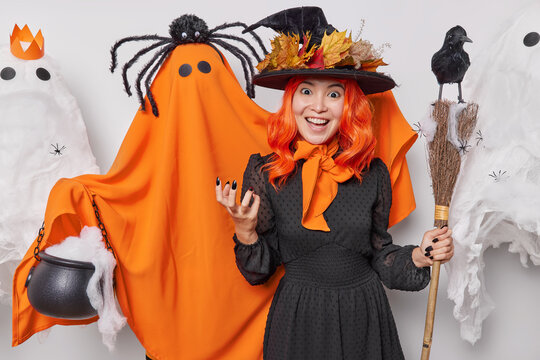 Holiday celebration concept. Indoor photo of young cheerful smiling European woman in costume of witch with broom organising Halloween party surrounded by scary ghosts looking straight at camera