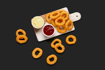 Board with fried breaded onion rings and different sauces on black background