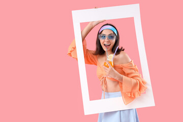Beautiful young woman with glass of tasty cocktail and frame on pink background