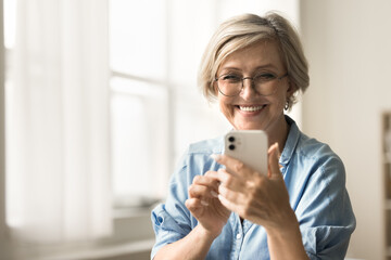 Positive trendy senior woman texting message on cellphone, taking selfie browsing Internet, social...