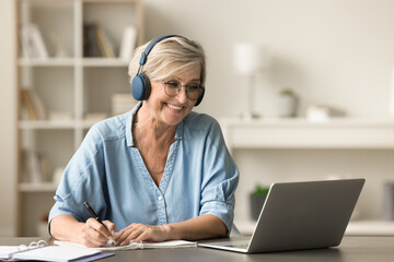 Happy senior retired student woman in earphones and glasses studying on Internet from home, writing...