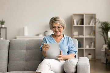 Happy pretty elder woman using digital tablet computer at home, reading electronic book, talking on...