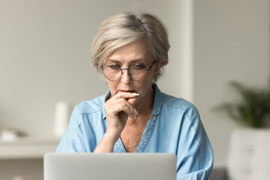 Focused senior business woman in elegant glasses thinking on work problem at laptop, touching chin, looking at laptop, making decision, using gadget for professional communication at home