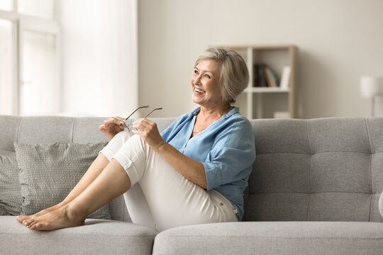 Happy Satisfied Old Lady Taking Glasses Off, Looking Away, Smiling, Laughing, Resting On Cozy Home Sofa. Elder Senior Patient Enjoying Good Vision, Strong Eyesight After Laser Eye Surgery