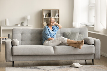 Cheerful dreamy senior pensioner woman relaxing on comfortable couch, outstretching barefooted...